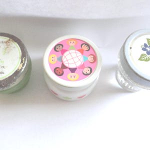 May include: Three vintage cosmetic jars with lids. One jar has a green base and a lid with text. Another jar has a pink lid with illustrated faces. The third jar has a clear glass base and a lid with a floral design.