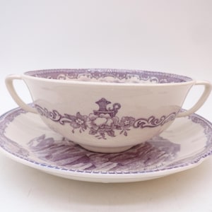 May include: Cream-colored soup bowl with two handles and matching saucer. Both pieces feature a purple floral and scroll design. The bowl sits on the saucer, creating a classic tableware set.