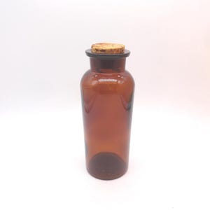 May include: A cylindrical amber glass bottle with a smooth, translucent finish and a wide neck. The bottle is topped with a light brown, textured natural cork stopper that fits securely into the opening, creating a simple and functional container.
