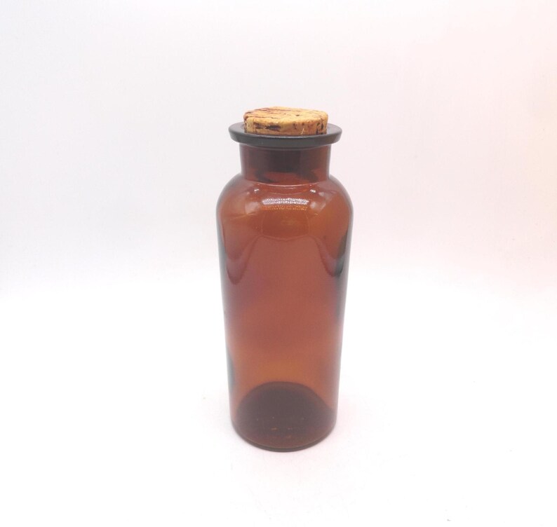May include: A cylindrical amber glass bottle with a smooth, translucent finish and a wide neck. The bottle is topped with a light brown, textured natural cork stopper that fits securely into the opening, creating a simple and functional container.