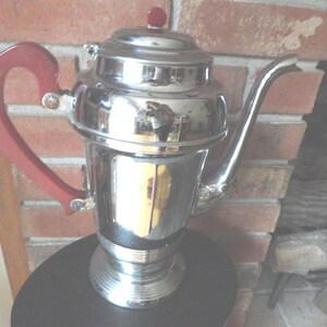 May include: A vintage chrome coffee pot with a red plastic handle and lid. The pot has a rounded body and a flared base.
