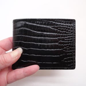 May include: A black bifold wallet featuring a glossy, embossed crocodile skin texture. The exterior surface displays a repeating scale pattern with a polished finish, showcasing a rectangular shape with clean, stitched edges along the perimeter.