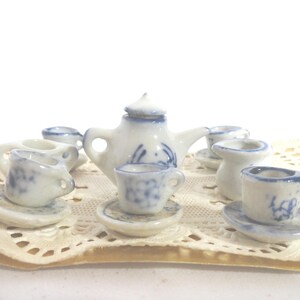 May include: Miniature porcelain tea set with a teapot, cups, and saucers. The set is white with blue floral designs. The tea set is displayed on a cream-colored lace doily.