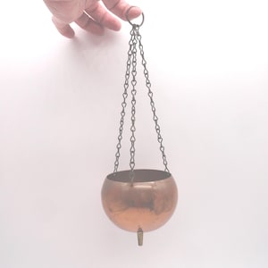 May include: A copper-colored hanging planter with a rounded bowl shape, suspended by three chains. The planter has a small, pointed foot at the bottom. The chains connect to a ring at the top.