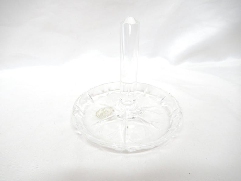 Op de afbeelding: Transparante glazen ringhouder met een ronde basis en een verticale paal. De basis heeft een decoratief, stervormig patroon. De ringhouder is gemaakt van helder glas. Het item staat op een witte achtergrond.