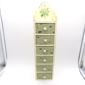 May include: A distressed, vintage-style wall-mounted cabinet with six drawers. The cabinet is painted in a distressed green and cream color scheme, with a floral design on the top. Each drawer has a white knob.