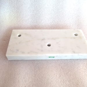 May include: A white marble shelf with three holes drilled into the top surface. The shelf is rectangular and has a smooth, polished finish.