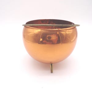 May include: A copper-colored, spherical bowl with a small, curved handle and three short legs. The bowl has a reflective surface and a dark interior, suggesting it may be an ashtray or decorative item.