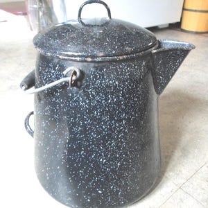May include: A black and white speckled enamelware coffee pot with a lid and a spout. The pot has a handle and a wire bail. The overall shape is a rounded cylinder. The pot is likely vintage.