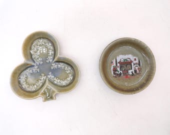 May include: A clover-shaped ceramic dish with a glossy olive-green glaze and white shamrock patterns, alongside a circular ceramic dish featuring a central illustration of a fireplace scene with a person sitting in a chair and a small dog.
