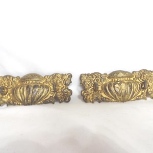 May include: Two antique gold-colored drawer pulls. Each pull features a central, urn-shaped design with a decorative lid, flanked by ornate, scrolling floral and foliate embellishments. The pulls are likely made of metal and show signs of age.
