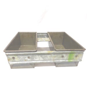 May include: A vintage metal bread pan with two rectangular compartments. The pan has a silver finish with visible wear and tear, and is designed to bake two loaves of bread simultaneously. The pan is set against a white background.