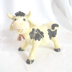May include: A ceramic cow figurine with a cream-colored body, black spots, and a copper bell. The cow has a smiling expression and black hooves. The figurine has a black patch of hair on its head.