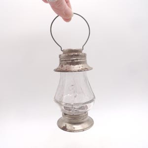 Pu&ograve; includere: Una piccola lanterna di vetro trasparente con base, parte superiore e manico in metallo. La lanterna ha un aspetto vintage e rustico con usura visibile sui componenti metallici. Il vetro &egrave; trasparente e il manico &egrave; un semplice anello di filo.