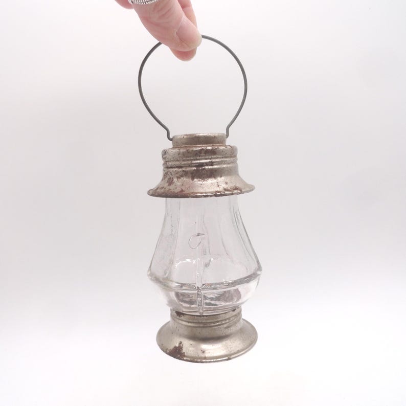 Pu&ograve; includere: Una piccola lanterna di vetro trasparente con base, parte superiore e manico in metallo. La lanterna ha un aspetto vintage e rustico con usura visibile sui componenti metallici. Il vetro &egrave; trasparente e il manico &egrave; un semplice anello di filo.