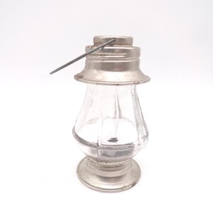 Pu&ograve; includere: Una piccola lanterna di vetro trasparente con coperchio, base e manico in metallo color argento. La lanterna ha una forma classica con un corpo arrotondato e una base svasata. I componenti metallici mostrano segni di et&agrave; e usura.