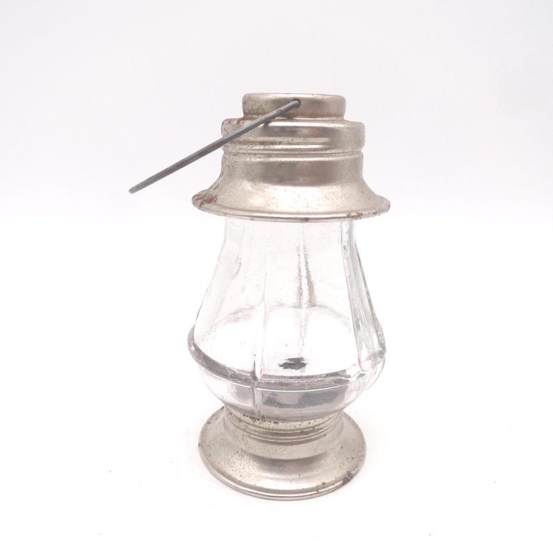 Pu&ograve; includere: Una piccola lanterna di vetro trasparente con coperchio, base e manico in metallo color argento. La lanterna ha una forma classica con un corpo arrotondato e una base svasata. I componenti metallici mostrano segni di et&agrave; e usura.