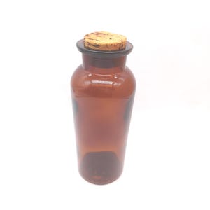 May include: A translucent amber glass bottle with a cylindrical body and a wide neck. The bottle is topped with a natural light brown cork stopper that features a textured, porous surface and dark brown grain patterns across the top.