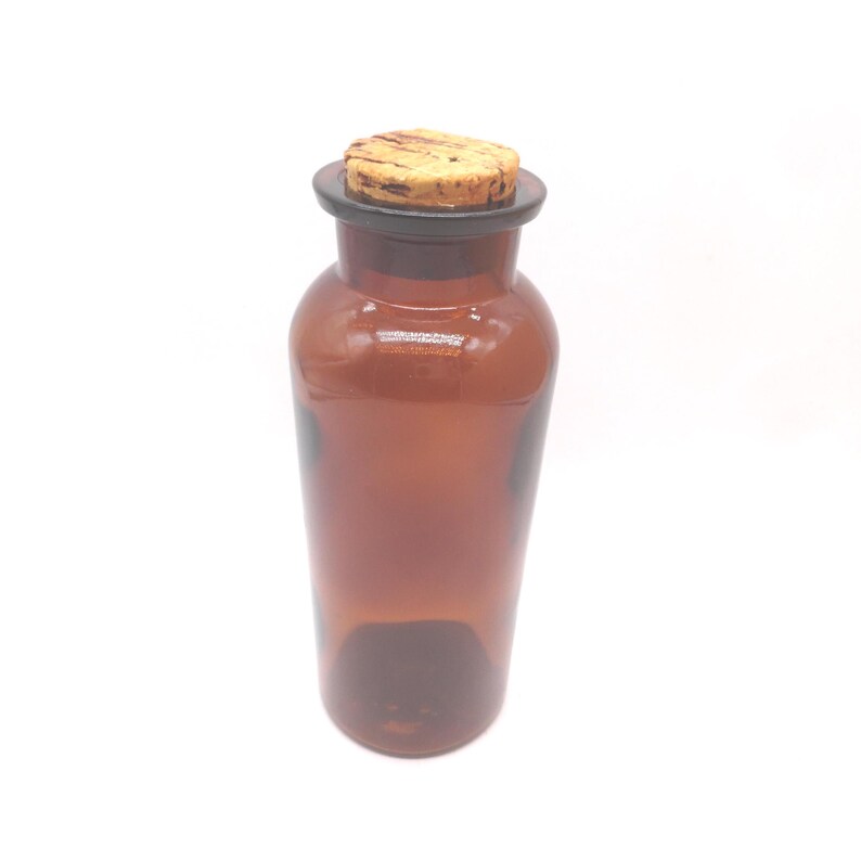 May include: A translucent amber glass bottle with a cylindrical body and a wide neck. The bottle is topped with a natural light brown cork stopper that features a textured, porous surface and dark brown grain patterns across the top.