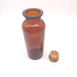 May include: A cylindrical amber glass bottle with a wide neck and a smooth, rounded rim. The bottle is translucent, allowing light to pass through the brown glass. Beside the bottle sits a small, round, light brown natural cork stopper.
