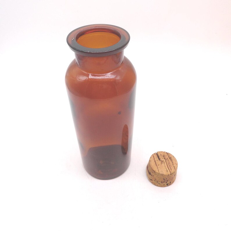 May include: A cylindrical amber glass bottle with a wide neck and a smooth, rounded rim. The bottle is translucent, allowing light to pass through the brown glass. Beside the bottle sits a small, round, light brown natural cork stopper.
