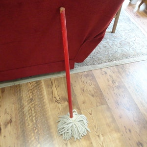 May include: A red mop handle with a white cotton mop head. The mop is standing on a wooden floor.
