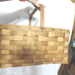 May include: A large, rectangular, woven wooden basket with a single handle. The basket is made of natural wood and has a light brown color. The basket is empty.