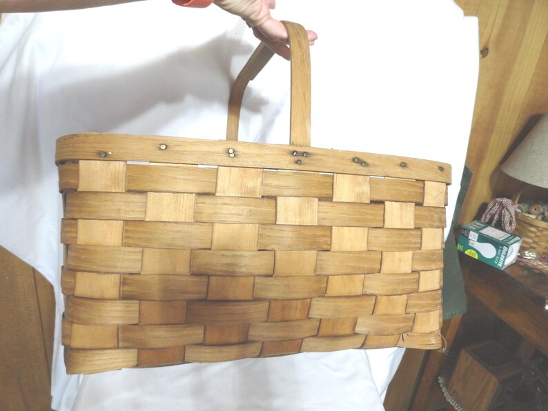 May include: A large, rectangular, woven wooden basket with a single handle. The basket is made of natural wood and has a light brown color. The basket is empty.