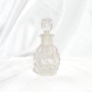 May include: A clear glass perfume bottle with a decorative stopper. The bottle has a textured, rounded body and a long neck. The stopper is also clear and has a detailed design. The bottle is empty and sits on a white surface.
