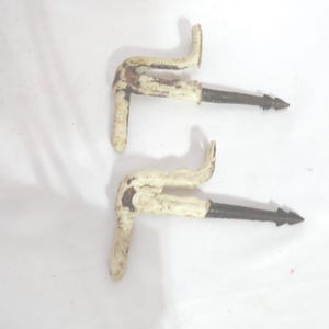 May include: Two antique, white-painted metal hooks with a curved handle and a long, pointed, dark-colored spike. The hooks show signs of wear and tear, suggesting they are vintage or reclaimed items.