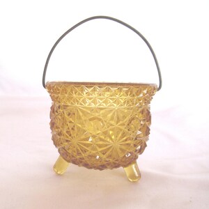 May include: A small amber glass cauldron with a wire handle and three legs. The cauldron has a diamond pattern.