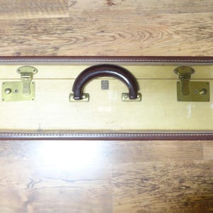 May include: A rectangular vintage suitcase featuring a cream-colored body with dark brown trim and a matching dark brown handle. The case includes two brass-toned metal latches and hinges, showing signs of wear on the exterior surface.