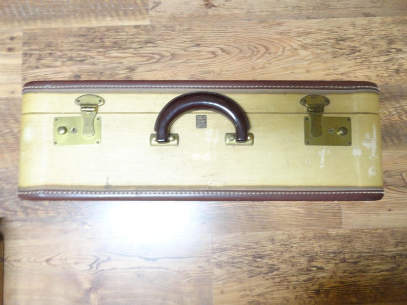 May include: A rectangular vintage suitcase featuring a cream-colored body with dark brown trim and a matching dark brown handle. The case includes two brass-toned metal latches and hinges, showing signs of wear on the exterior surface.