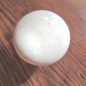 May include: A spherical white ceramic knob with a smooth, glossy finish and subtle speckled texture. The object is round and features a uniform, bright white surface that reflects light, creating a clean and simple appearance for furniture hardware.