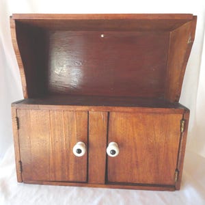May include: A wooden cabinet with two doors and a top shelf. The cabinet is made of dark brown wood and has white and black knobs. The doors are closed, and the cabinet is empty. The cabinet is likely used for storage.