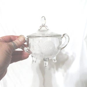 May include: Clear glass sugar bowl with a lid, handles, and three feet. The bowl features an etched floral design. The lid has a small handle. The sugar bowl is held by a hand.