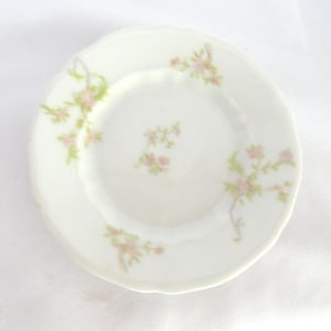 May include: A small, white porcelain plate with a scalloped edge. Delicate pink and green floral sprigs are arranged around the plate's rim and center, creating a vintage aesthetic. The plate is likely for serving small portions.