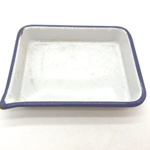 May include: A square enamel tray with a white interior and a dark blue rim. The tray features a small, pointed pouring spout on one corner. The surface shows signs of wear and minor discoloration, consistent with a vintage metal kitchen item.