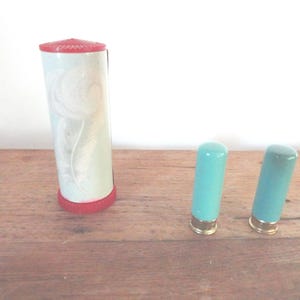 Puede incluir: Un recipiente alto y cilíndrico con tapa y base rojas, y un cuerpo azul claro con un diseño floral. También hay dos objetos pequeños de color turquesa con bases doradas. Los artículos están sobre una superficie de madera.