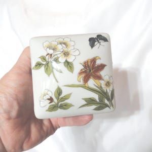 May include: Square porcelain trinket box with a white background and floral design. The box features white, yellow, brown, and green flowers, as well as a black butterfly.