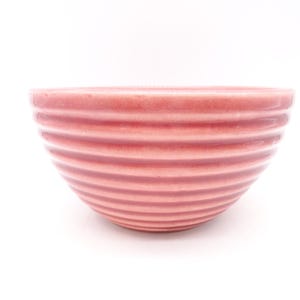 May include: A pink ceramic bowl with a ribbed design. The bowl is a soft, muted pink color and has a smooth, glossy finish. The horizontal ridges create a textured appearance. The bowl is set against a white background.