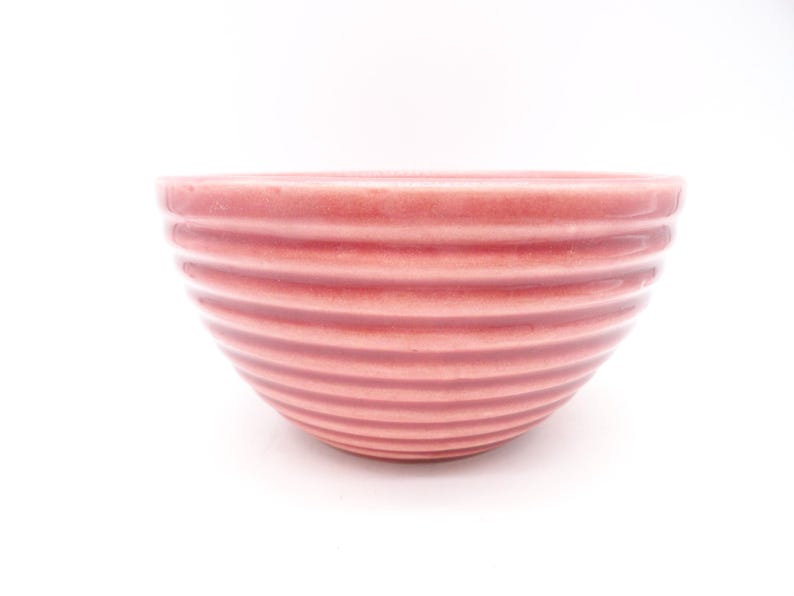 May include: A pink ceramic bowl with a ribbed design. The bowl is a soft, muted pink color and has a smooth, glossy finish. The horizontal ridges create a textured appearance. The bowl is set against a white background.