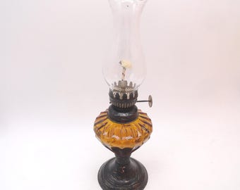 May include: An oil lamp featuring a clear glass chimney with a ruffled top, a dark metal burner assembly with a side adjustment knob, a textured amber glass reservoir with vertical ribbing, and a dark, circular metal base with embossed details.