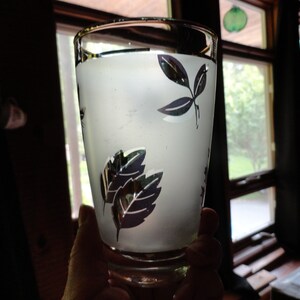 May include: A frosted glass with a black and white leaf design, filled with a clear liquid. The glass is being held by a hand.