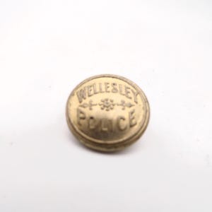 May include: A round, antique brass-colored button with the words "WELLESLEY POLICE" embossed on the surface. The text is arranged in a circular pattern, with a decorative emblem between the words.