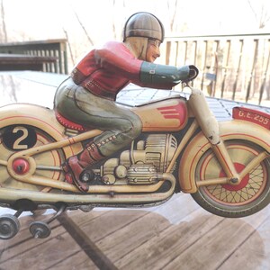 May include: A vintage tin toy motorcycle with a rider wearing a helmet and goggles. The motorcycle is painted in cream, red, and green with a number "2" on the side. The motorcycle has a wind-up key and is labeled "G.E. 255".