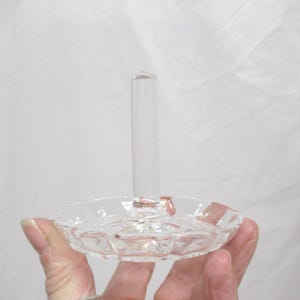 Op de afbeelding: Een heldere glazen ringhouder met een ronde, facetgeslepen basis en een lange, cilindrische paal. De ringhouder wordt in de hand van een persoon gehouden tegen een witte achtergrond. De ringhouder is ontworpen om ringen te bevatten.