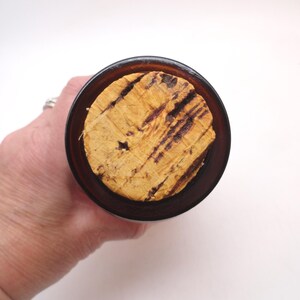 May include: A top-down view of a cylindrical amber glass bottle opening sealed with a natural, light-tan cork stopper. The cork features a textured, porous surface with dark brown, linear grain patterns running across its circular top face.