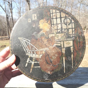 May include: Round, vintage tin with a black background and a detailed illustration. The illustration depicts a person seated at a desk near a window. The person is wearing a brown dress and sitting in a white chair.
