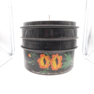 May include: A black metal container with three stacked sections. The bottom section features a hand-painted design of orange and yellow flowers with green leaves. The container has a lid and a small handle on top.
